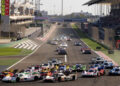 Racing cars, including Porsche models driven by skilled works drivers, compete on a track in bright daylight. Spectators fill the stands beneath a large FIA WEC 2024 banner overhead.