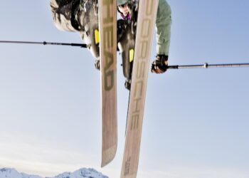 A skier executes a mid-air trick against a clear blue sky, with snow-covered mountains in the background, showcasing precision akin to sports car engineering.