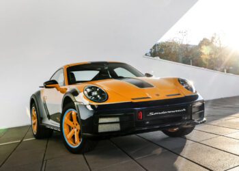 The one-off black and orange Porsche 911 Dakar is elegantly parked on a tiled surface, with a sleek modern building rising majestically in the background.
