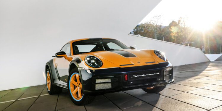 The one-off black and orange Porsche 911 Dakar is elegantly parked on a tiled surface, with a sleek modern building rising majestically in the background.