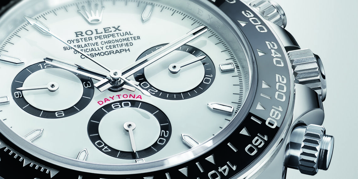 Close-up of the ultimate Rolex Daytona chronograph, featuring a white face, black ceramic bezel, and three sub-dials displaying hours, minutes, and seconds. The word "Daytona" is in red above the lower sub-dial, embodying its iconic automotive watch heritage.