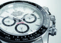 Close-up of the ultimate Rolex Daytona chronograph, featuring a white face, black ceramic bezel, and three sub-dials displaying hours, minutes, and seconds. The word "Daytona" is in red above the lower sub-dial, embodying its iconic automotive watch heritage.