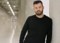 A man with a beard, resembling Mate Rimac, leans against a concrete wall in a well-lit hallway. Dressed in a black sweater and grey pants, he stands confidently with hands in pockets, embodying the innovative spirit of the Rimac Group CEO.