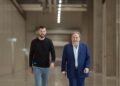 Two men walking in a spacious industrial hallway with overhead lighting. One, dressed in a black shirt and jeans, is a close associate of Mate Rimac, the visionary behind Rimac Group. The other, in a sharp blue suit, seems poised to discuss the latest news about their CEO stepping down.