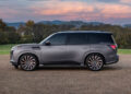 A sleek, matte gray Infiniti SUV is parked on a gravel path, exuding tailgating luxury against a backdrop of trees and a cloudy sky at sunset.