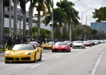 A line of exotic sports cars, including yellow and red ones, speeds down a palm-lined street in South Florida, with eager spectators lining the sidewalk for this thrilling toy rally.