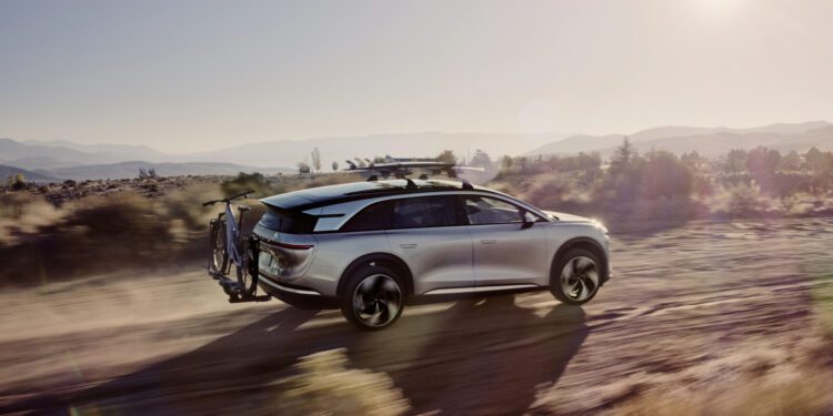A silver SUV with a roof rack and a bicycle mounted on the rear drives on a dirt road in a desert landscape during sunset.