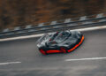 A black and red Bugatti W16 Mistral blazes down the curving road at blistering speeds, its sleek open-top design cutting through the air with precision. The blurred background highlights its relentless pursuit of setting an open-top speed record.