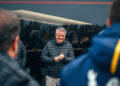 A smiling man in a black jacket stands outside, surrounded by others in jackets, as they all admire the sleek lines of a Bugatti W16 Mistral.