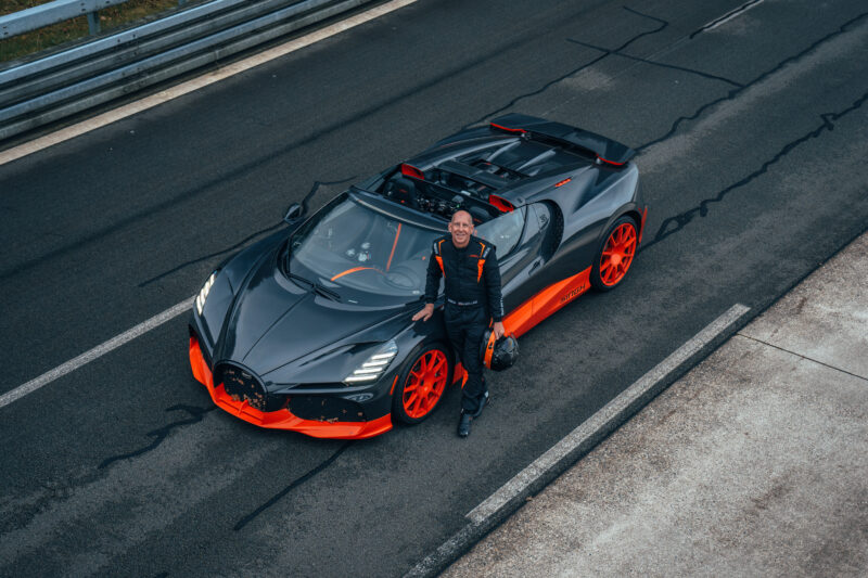 A person in racing gear stands next to a sleek, black and orange Bugatti on the racetrack, ready to chase the speed record with the powerful W16 Mistral.