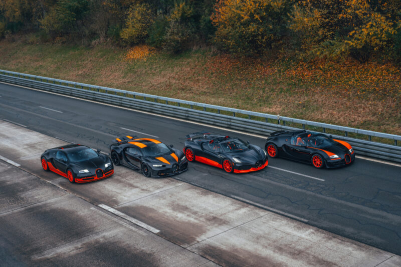 Four high-performance sports cars, including the Bugatti W16 Mistral known for its open-top speed record of 282.05 MPH, are parked in a line on a paved road, surrounded by trees with autumn foliage.