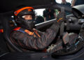 A person wearing a racing suit and helmet sits confidently in the driver's seat of a Bugatti, hands gripping the steering wheel, ready to chase the speed record in their high-performance W16 Mistral sports car.