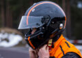 A person wearing an orange and black helmet, reminiscent of the sleek Bugatti style, adjusts the strap while dressed in an orange suit. Snow and trees frame the scene, capturing the essence of speed and precision.