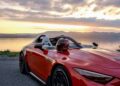 A red Mercedes-AMG convertible sports car, embodying PureSpeed, is parked by the lakeside at sunset, with a helmet resting on the passenger seat.