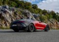 The Mercedes-AMG convertible sports car, a PureSpeed marvel in red and black, is parked on a road beside rocky cliffs and lush greenery under a partly cloudy sky.