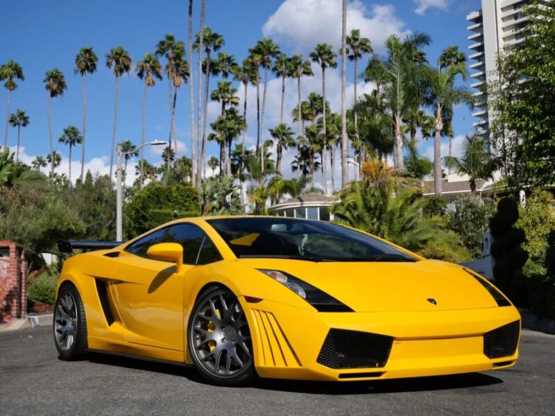 A yellow Lamborghini Gallardo is parked on the street, flanked by palm trees and buildings under a partly cloudy sky.