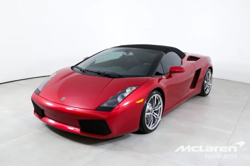 A red convertible sports car with a black roof is parked indoors on a light-colored floor. The car boasts a sleek design, silver wheels, and embodies the allure of a V10 supercar. "McLaren Charlotte" is visible in the bottom right corner, hinting it's for sale among other elite automobiles.