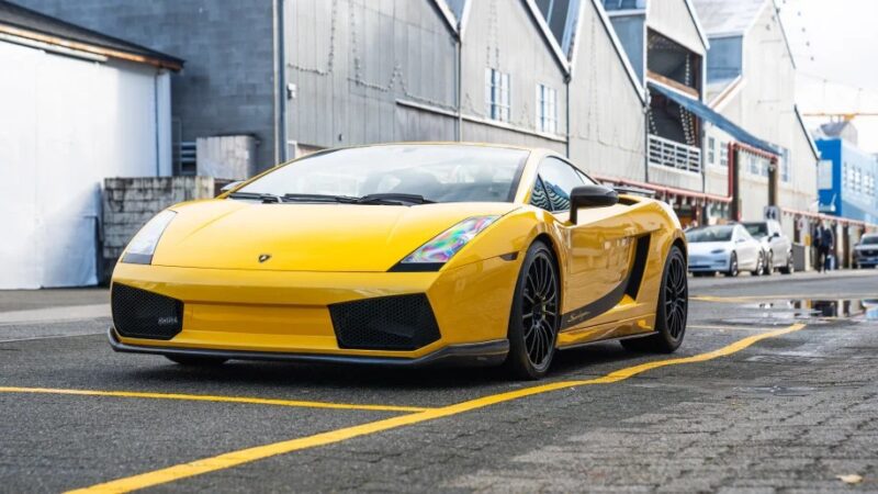 A sleek yellow supercar, possibly one of those impressive Lamborghini Gallardos, is parked on a street with industrial buildings looming in the background.