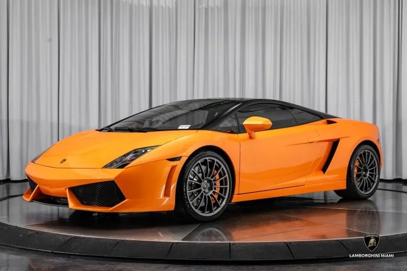 Orange Lamborghini Gallardo with black roof on display platform, showcasing its V10 supercar prowess in a showroom with white curtains. This stunning Lamborghini Gallardo is now for sale, offering an unparalleled blend of style and performance.