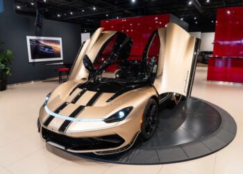 A gold and black electric hypercar with its scissor doors open is displayed in a showroom with a red and black backdrop, highlighting the stunning Pininfarina Battista for sale.