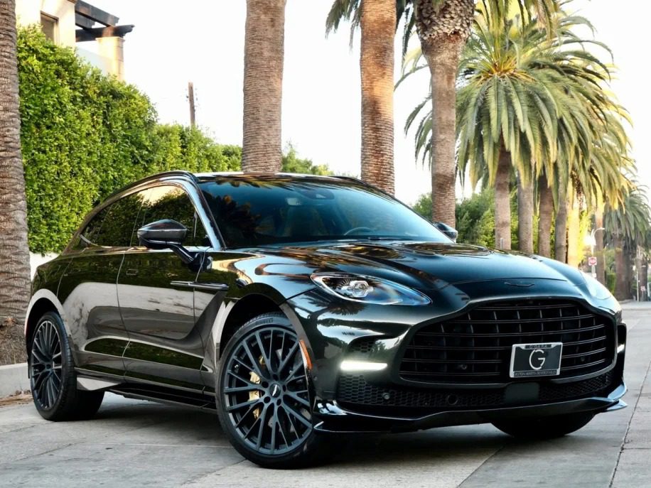 A sleek, black Aston Martin DBX 707 luxury SUV is parked on a sunny street lined with palm trees, exemplifying the allure of Super-SUVs, and it's for sale.