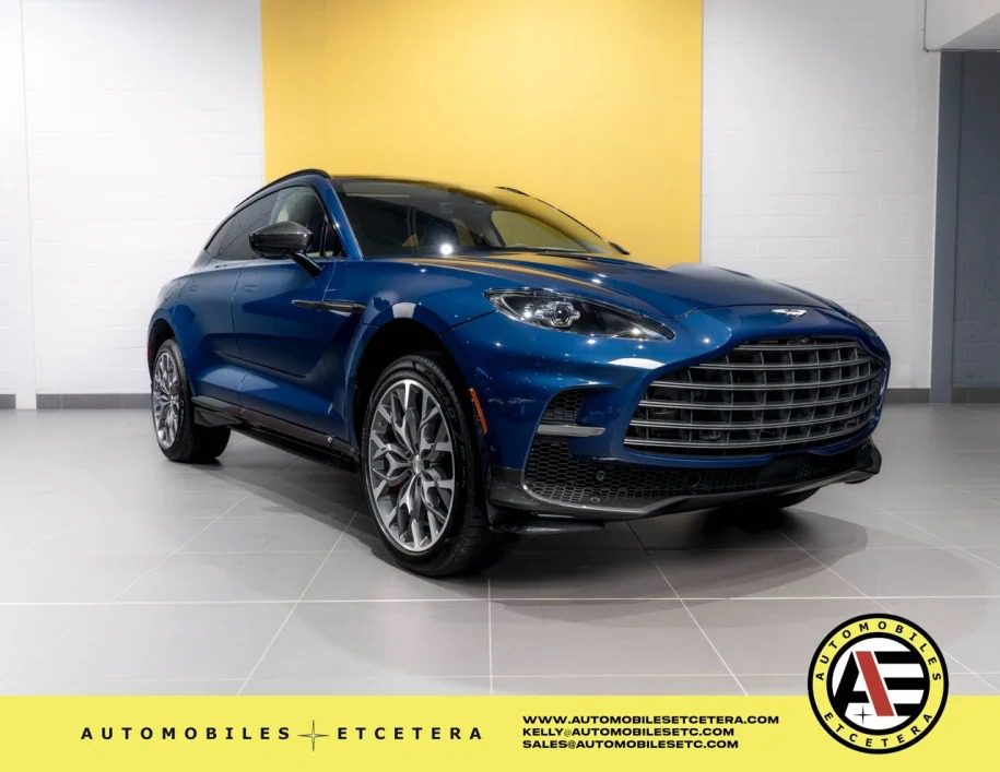 A blue Aston Martin DBX 707, one of the finest super-SUVs, is parked indoors on a tiled floor. A yellow and white wall serves as the backdrop while the brand logo is tastefully displayed in the bottom right corner, signaling its prestige and availability for sale.