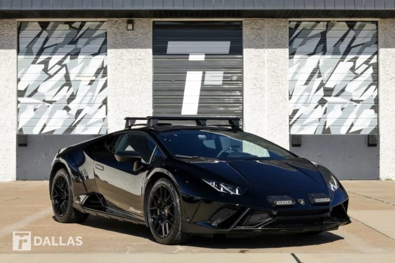 A stunning black Lamborghini Huracán Sterrato with a roof rack is elegantly parked in front of a building, its garage doors boasting eye-catching geometric patterns.