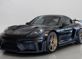A sleek black Porsche 718 Cayman GT4 RS with gold rims and a rear spoiler is on display in the showroom. Track-ready and stunning, this sports car is now for sale.
