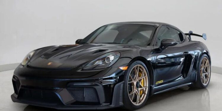 A sleek black Porsche 718 Cayman GT4 RS with gold rims and a rear spoiler is on display in the showroom. Track-ready and stunning, this sports car is now for sale.