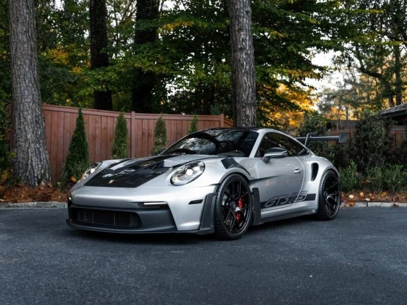 A silver Porsche 911 GT3 RS with black accents is parked on a tree-lined street, showcasing its distinct rear wing. Known as the ultimate driver's car, this sports vehicle proudly displays its GT3 RS signage.