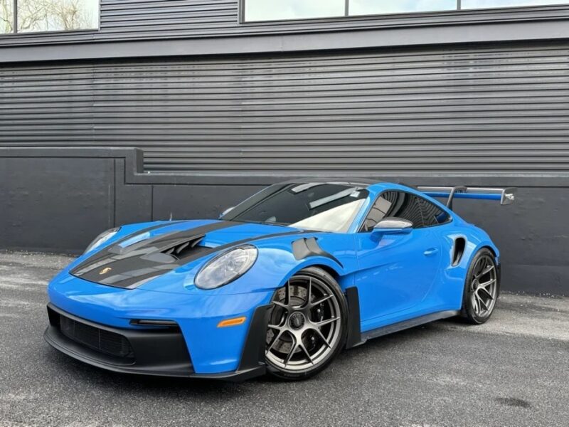 A sleek blue sports car, reminiscent of a Porsche 911 GT3 RS with its black hood and large spoiler, is parked in front of a modern, dark gray building.