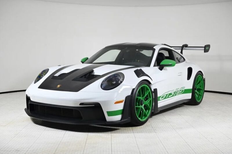 A white Porsche 911 GT3 RS with green accents, large rear wing, and matching green wheels is showcased in a well-lit showroom. This ultimate driver's car radiates an aura of precision and power, captivating all who gaze upon it.