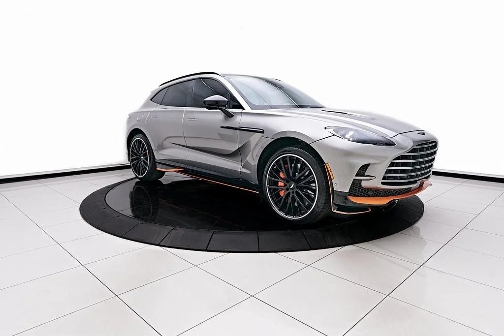 A stunning Silver luxury SUV, the Aston Martin DBX 707, graces a rotating display platform. With its black and orange accents, this super-SUV is showcased in a brightly lit room with a tiled floor. For sale now, it promises unparalleled luxury and performance.