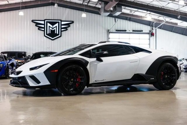 A white Lamborghini, possibly a Huracán Sterrato, with a sleek design is parked indoors. The sports car features black accents and tinted windows, surrounded by other vehicles. A logo with the letter "M" graces the wall, hinting at its prestigious surroundings and tempting any onlooker to consider a Lamborghini purchase.