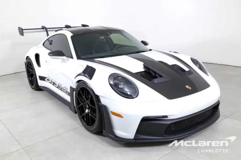 White Porsche 911 GT3 RS with black accents and a large rear wing, the ultimate driver's car, elegantly poised on a light gray floor. Logo in the corner reads "McLaren Charlotte.