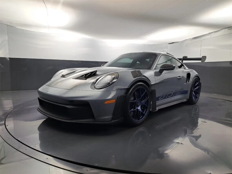 In the showroom under bright lights, a sleek silver Porsche 911 GT3 RS stands out with its large rear wing, blue wheels, and "GT3 RS" decals. Truly a driver's car, this masterpiece exemplifies precision and performance.