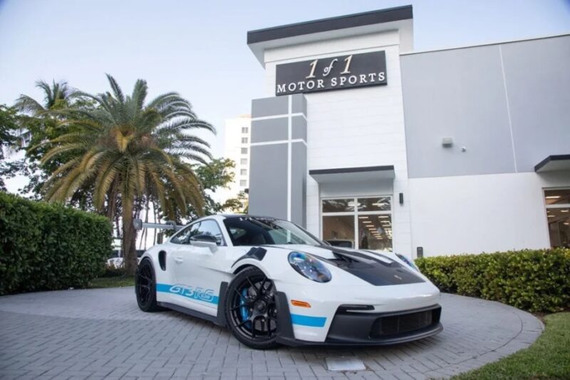 A Porsche 911 GT3 RS, the ultimate driver's car, is parked in front of "1 of 1 Motor Sports," with palm trees swaying in the background.