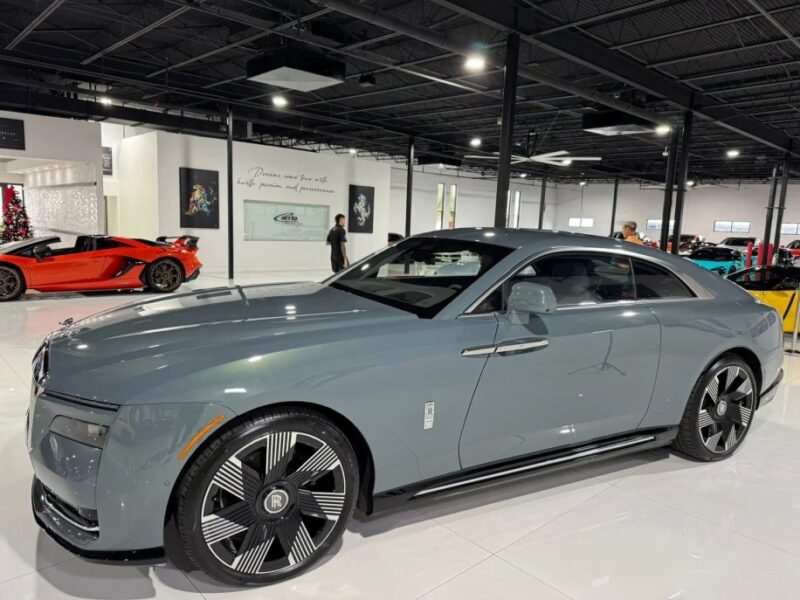 A gray Rolls-Royce Spectre, an epitome of ultra-luxe design, sits gracefully indoors in a showroom. Behind it, a vibrant red sports car adds contrast while people admire these marvels of engineering.