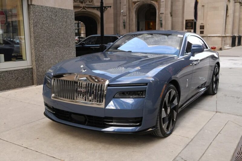 A sleek, blue Rolls-Royce Electric Super Coupe is parked on a city street in front of a stone building, showcasing its shiny grille and modern design.