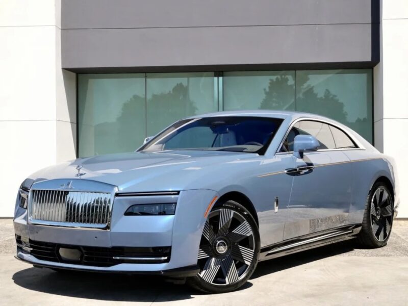 The Electric Ultra-Luxe silver super coupe boasts a distinctive front grille and sleek design, exuding elegance. Parked on a concrete surface, it sits in front of a building with large windows—a testament to the modern luxury of the Rolls-Royce Spectre.