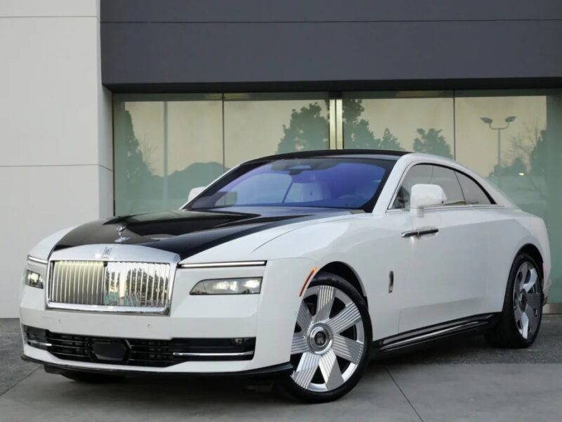 A white Electric Ultra-Luxe coupe with a black hood is elegantly parked in front of a modern building, possibly hinting at the sophistication of the Rolls-Royce Spectre.