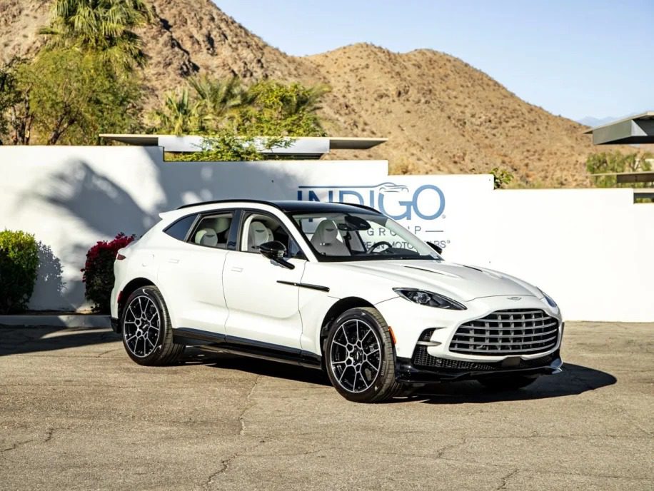 A white luxury Aston Martin DBX 707, part of the elite Super-SUVs lineup, is elegantly parked on the pavement. In the background, desert mountains rise majestically alongside an "Indigo" sign on a pristine white wall, creating an alluring backdrop for this beautiful vehicle.