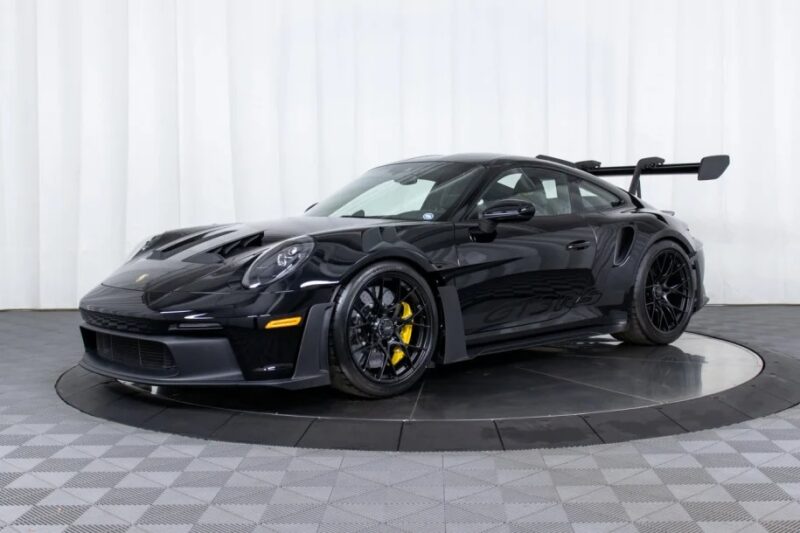 A Porsche 911 GT3 RS, known as Porsche's ultimate driver's car, stands proudly in the showroom. Its sleek black design and large rear wing contrast strikingly against the checkered floor and white backdrop.