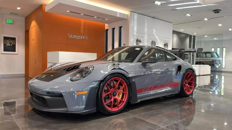 A gray Porsche 911 GT3 RS with red rims and the "GP RS" label stands proudly in a modern showroom. Its sleek design epitomizes the ultimate driver's car, perfectly accented by shiny floors and an orange accent wall.