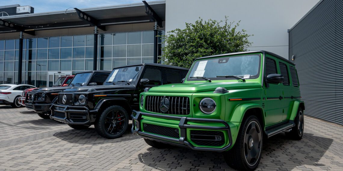 Several black and one green luxury SUVs are parked outside a modern Mercedes-Benz dealership building on a paved area.