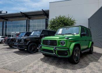 Several black and one green luxury SUVs are parked outside a modern Mercedes-Benz dealership building on a paved area.