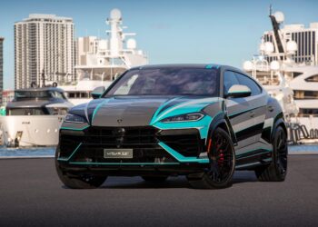 A black and turquoise Lamborghini Urus SE luxury SUV is parked near a marina, complemented by yachts and high-rise buildings in the background under a clear blue sky.
