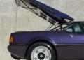 A purple BMW M1 sports car with its hood open, showcasing part of the rear and tire against a concrete wall backdrop, exuding the signature style of KITH and Ronnie Fieg.