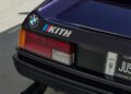 Rear view of a BMW M1 car with a distinctive KITH decal on the trunk lid, designed by Ronnie Fieg, showcasing its classic taillight design and part of a custom license plate.