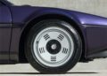 Close-up of a car wheel with a silver rim featuring a unique design of slotted holes, set against a sleek, dark purple car body, reminiscent of the stylish collaborations like KITH x BMW envisioned by founder Ronnie Fieg.